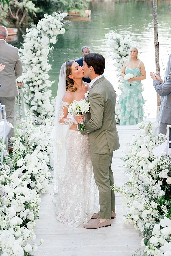 Wedding kiss on a wooden dock, bride in lace dress and veil with bouquet, groom in suit beneath a white rose arch by the lake