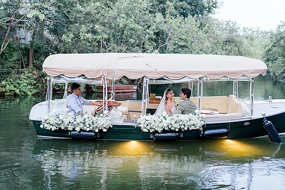 Couple portrait of newlyweds on a boat, bride in veil holding a white bouquet beside groom in a light suit on a river with trees