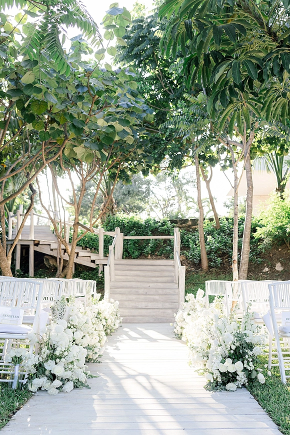 Ceremony aisle decor with white aisle flowers and greenery lining a wooden walkway, reserved white chairs set in a sunlit garden setting
