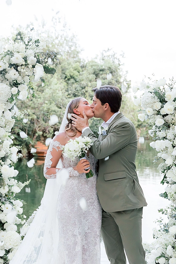 Wedding kiss portrait of bride and groom kissing under a white rose floral arch, long veil and bouquet, with lakeside trees and reflections behind