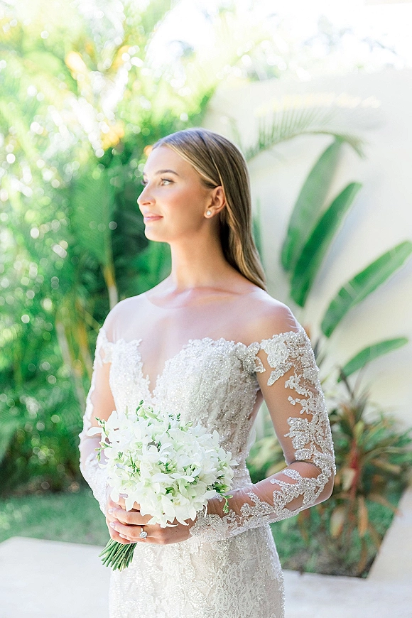 Bridal portrait of a bride holding bouquet in an off-the-shoulder lace long sleeve gown, standing in sunlight by tropical palm greenery