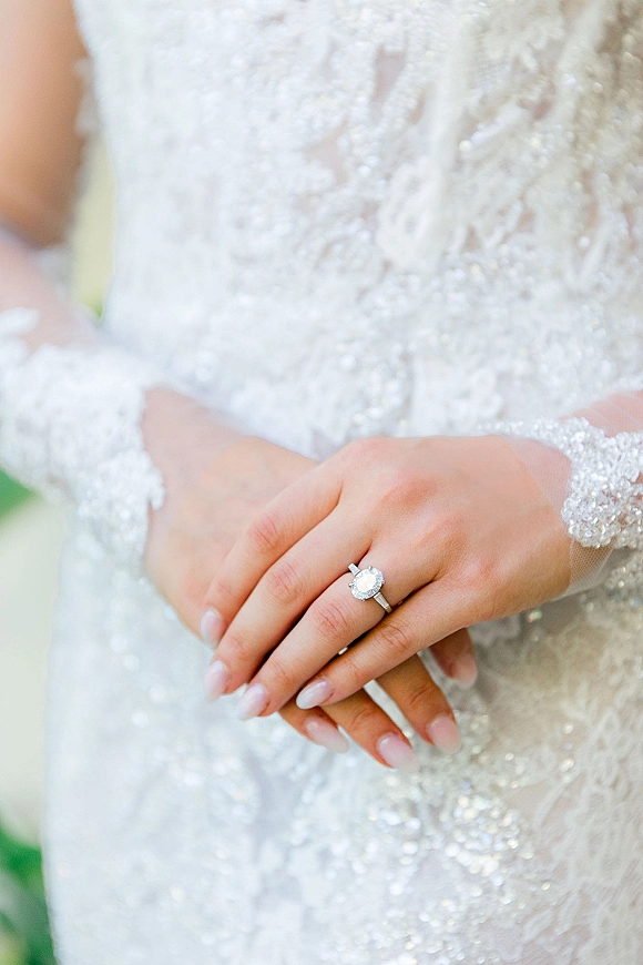 Engagement ring with a halo engagement ring design on a bride’s manicured hand, framed by lace sleeves and a softly blurred background