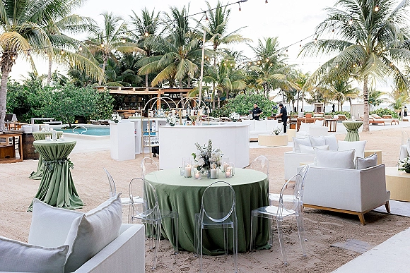 Beach reception decor with sage green tablecloths on round tables, clear acrylic chairs and white sofas on sand by palms and ocean view
