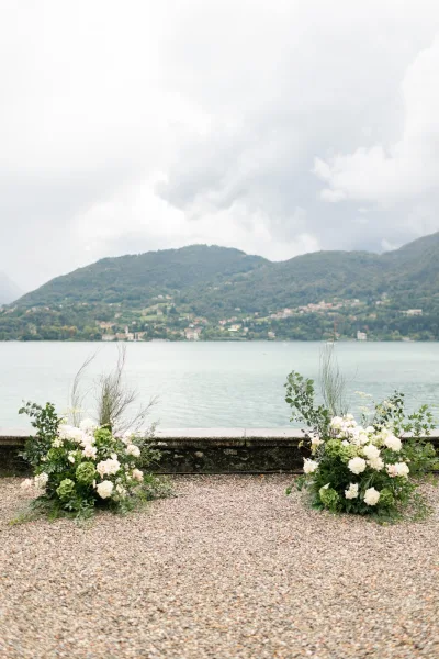 Ceremony floral arrangements with wedding aisle flowers of white roses, hydrangeas, and greenery by a stone wall with lake and mountains beyond