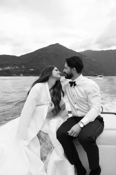 Couple portrait in a black and white wedding portrait style, newlyweds holding hands on a boat with mountains and a cloudy lake backdrop