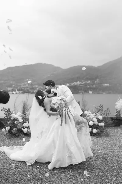 Wedding kiss portrait of bride and groom in a dip kiss by a lakeside mountain shoreline, bride in lace gown with veil and bouquet amid rose petals