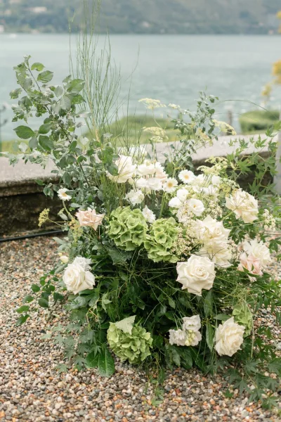 Wedding florals ground floral arrangement of white and blush roses with green hydrangea on gravel by a stone wall near a lake