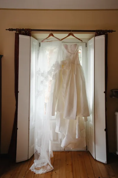 Wedding dress on hanger, strapless wedding dress glowing in window light with a lace veil, framed by white shutters and wood floor