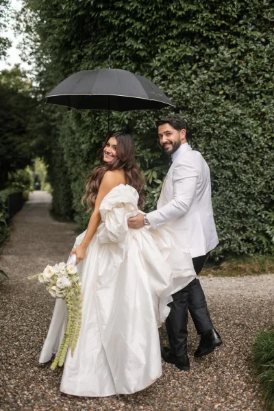 Couple portrait in rainy wedding photos, bride looking back as groom walks with her under a black umbrella along a garden hedge path