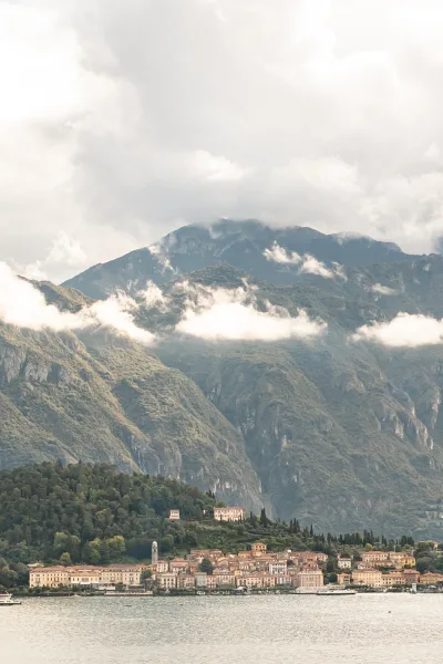 Lake mountain view with pastel lakeside village and church bell tower, boats on calm water beneath misty clouds and alpine peaks