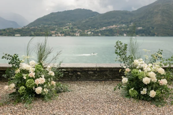 Ceremony altar flowers with white and blush roses and green hydrangea clusters along a gravel aisle, set by a lakeside with mountains beyond