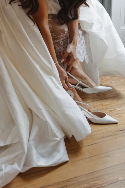 Bridal shoes, white wedding heels with pointed toes and ankle straps beside lace lingerie and bracelet on a wood floor in window light