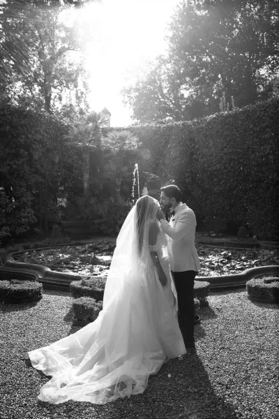 Couple portrait with flowing wedding veil train as groom in white tuxedo kisses bride’s forehead beside a sunlit garden fountain pond