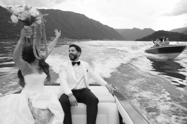 Wedding boat portrait of newlyweds on a speedboat, bride holding bouquet overhead beside groom in white tuxedo on a mountain lake