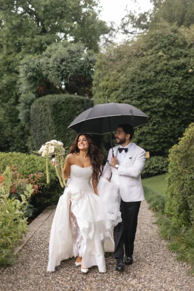 Couple portrait of bride and groom under umbrella, walking on a gravel garden path; she holds a bouquet as he wears a white tuxedo jacket.