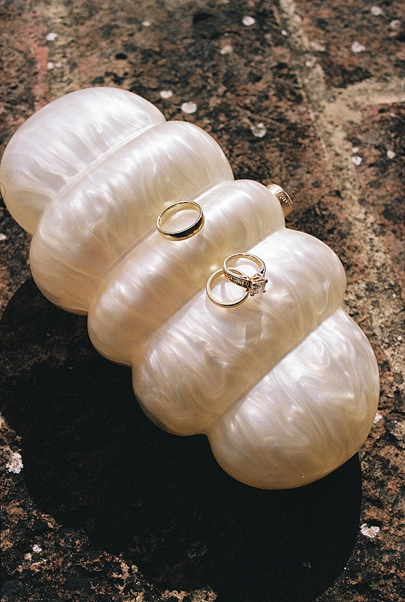 Wedding rings arranged in a wedding ring flat lay on a pearl clutch, featuring gold bands and a diamond engagement ring on sunlit stone