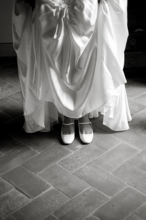 Bridal shoes, white wedding shoes in Mary Jane style tucked under a bridal gown with tulle underskirt on stone tile by a dresser