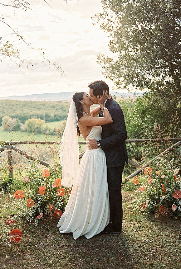Wedding kiss portrait of bride and groom kissing, her long veil flowing beside a wooden fence with rolling hills in the background
