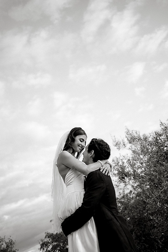 Couple portrait in a black and white wedding portrait, groom lifting bride as they embrace under a cloudy sky with trees behind