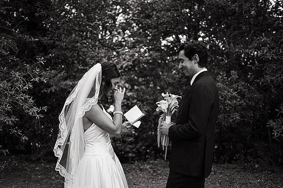 Wedding vows as bride reading vows to groom holding bouquet, lace-edge veil accent, in a garden ceremony with trees and lawn backdrop
