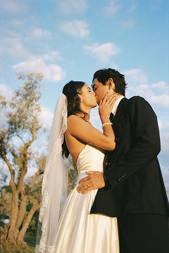 Wedding kiss as bride and groom kissing in side profile, her long veil flowing against a blue sky with clouds and trees behind