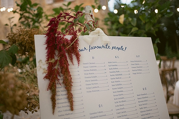 Wedding seating chart on an easel with a ribbon bow, hanging red florals and greenery under bistro string lights at an outdoor reception setting