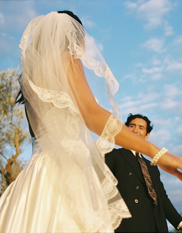 Couple portrait of bride and groom holding hands as her lace-trim veil blows, framed by blue sky, soft clouds, and trees