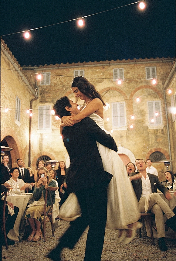 Wedding reception dance as groom lifts bride in satin dress under string lights in a courtyard, guests cheering by stone arches at night