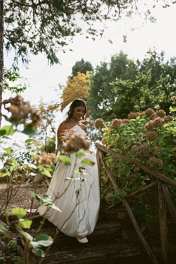 Bridal portrait of a bride walking down wooden garden steps, looking down in a one-shoulder satin dress with veil and rose bouquet