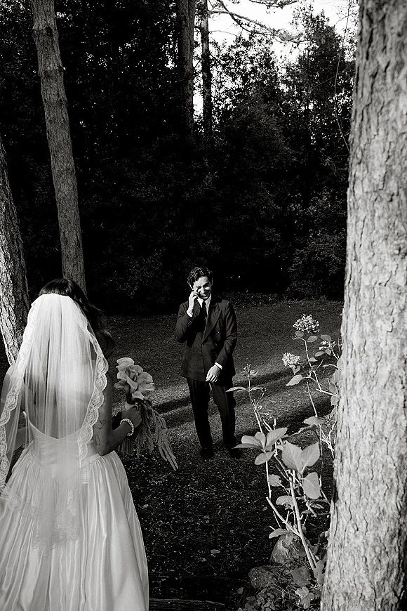 Wedding first look as bride walks up behind groom in a dark suit, bouquet in hand, veil trailing on a garden lawn among trees