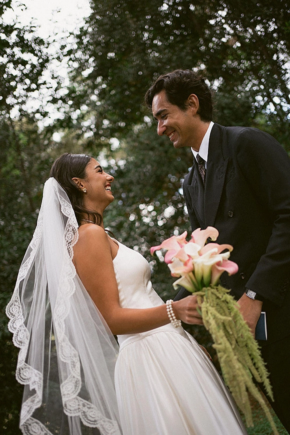 Couple portrait of bride and groom laughing, holding hands as she looks up at him, with lace veil and calla lily bouquet in a garden setting