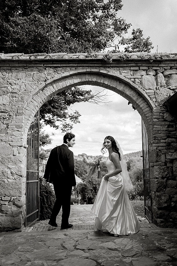 Couple portrait in a black and white wedding photo, bride in veil with bouquet and groom in tuxedo under a stone archway gate