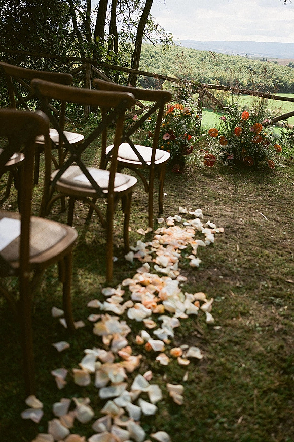 Ceremony aisle decor with a flower petal aisle and white runner, lined by wooden chairs and wildflowers on a hillside overlook with trees and mountains