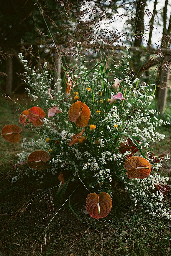 Wedding floral arrangement with anthurium and calla lilies, baby's breath and billy balls arranged low on grass beside forest trees
