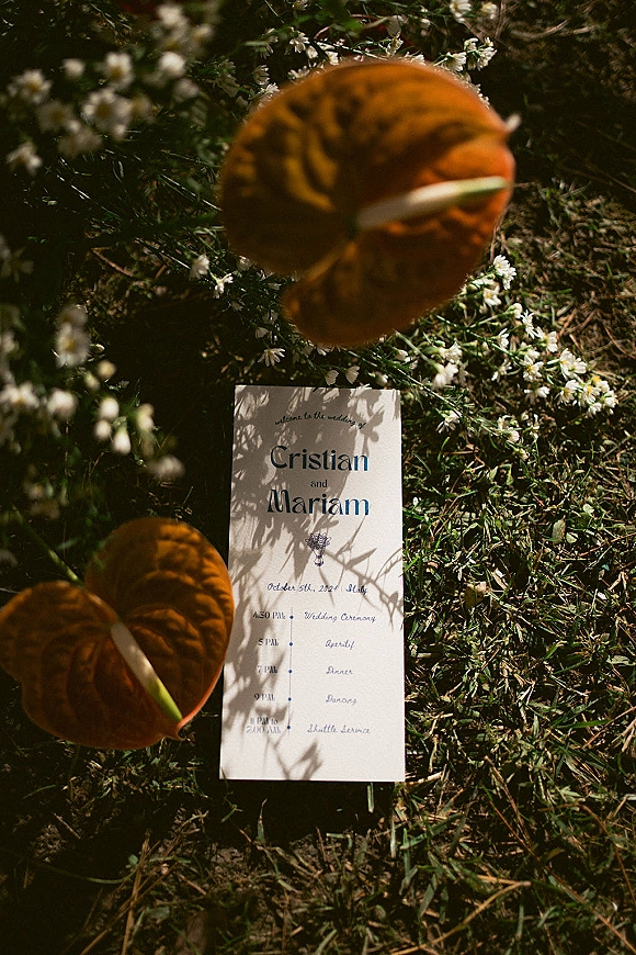 Wedding invitation flat lay with calla lilies and ribbon on grass, surrounded by small white flowers and dappled sunlight shadows