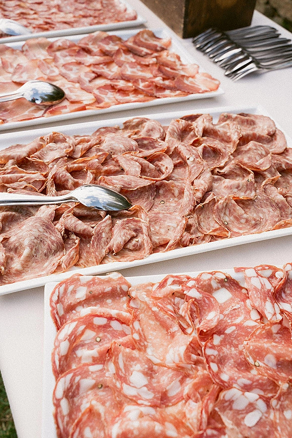 Charcuterie platter with sliced salami and cured meats arranged on white serving platters with napkins and utensils on a buffet table