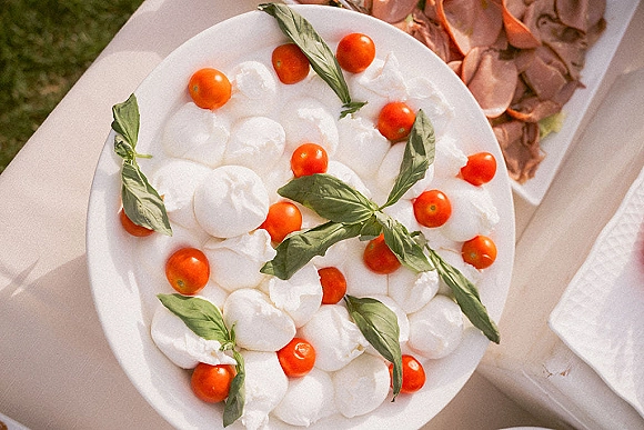 Caprese salad platter with wedding cocktail hour food featuring fresh mozzarella, cherry tomatoes, and basil on a white platter on tablecloth