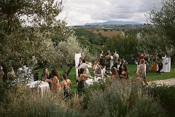 Wedding cocktail hour with outdoor wedding cocktail hour tables on a garden lawn, white linens, cocktails, floral centerpieces, olive trees and hills backdrop