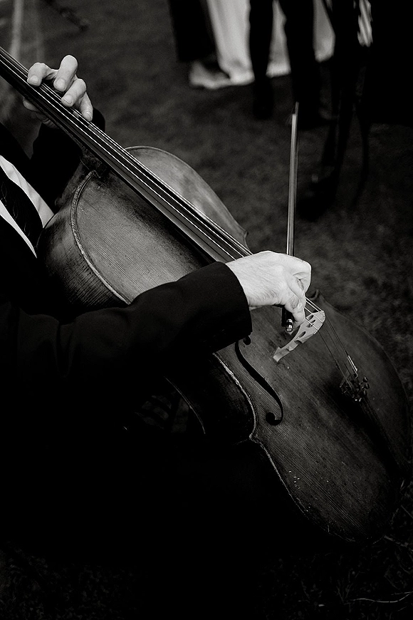 Wedding ceremony music as a cellist’s hands guide the bow across a cello, suit jacket visible, with guests standing on a grassy lawn