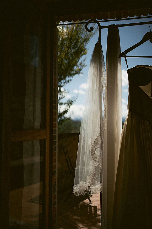 Wedding veil with lace accents hanging on a dress hanger in window light, softly backlit with mountains and patio tiles outside
