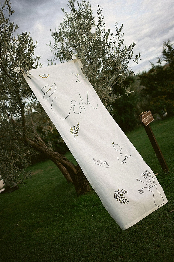 Wedding welcome sign on fabric with calligraphy lettering and botanical line drawings, hanging from a clip outdoors under tree branches