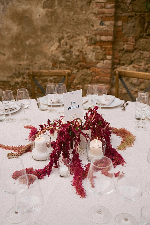 Reception tablescape with a burgundy wedding table centerpiece, glass hurricane candles, and place settings on white cloth against rustic brick wall