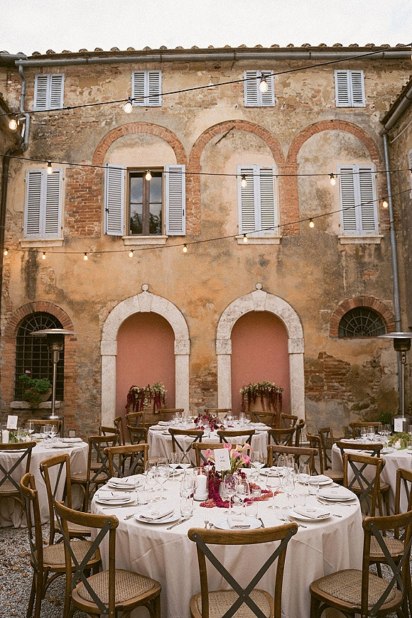 Reception tablescape for an outdoor wedding reception with round white-linen tables, candles and floral centerpieces under string lights in a rustic courtyard
