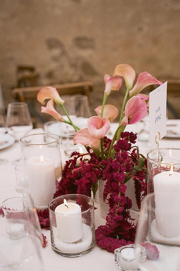 Reception tablescape with wedding table centerpiece of pink calla lilies, pillar and votive candles, wine glasses, and menu cards by a stone wall