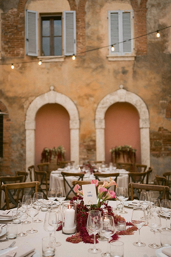 Reception tablescape with a round wedding table setup, white tablecloth, calla lily centerpiece, candles in hurricanes, and string lights in a stone courtyard