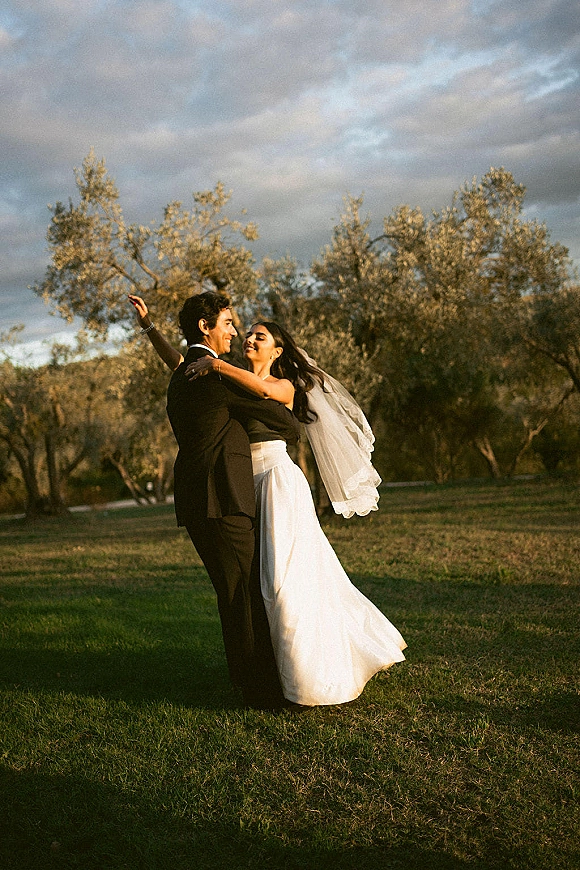 Couple portrait of groom holding bride in a wedding dip, veil blowing over strapless dress and tuxedo in a grassy park field