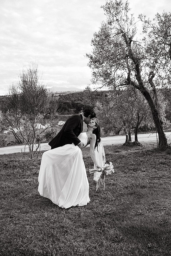 Wedding kiss portrait with a dip kiss wedding photo as groom dips bride in strapless dress and veil, bouquet in hand in a grassy field under cloudy sky