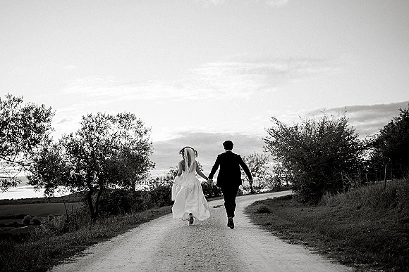 Couple portrait of newlyweds holding hands walking away down a dirt road, bride’s long veil and flowing dress train in countryside﻿