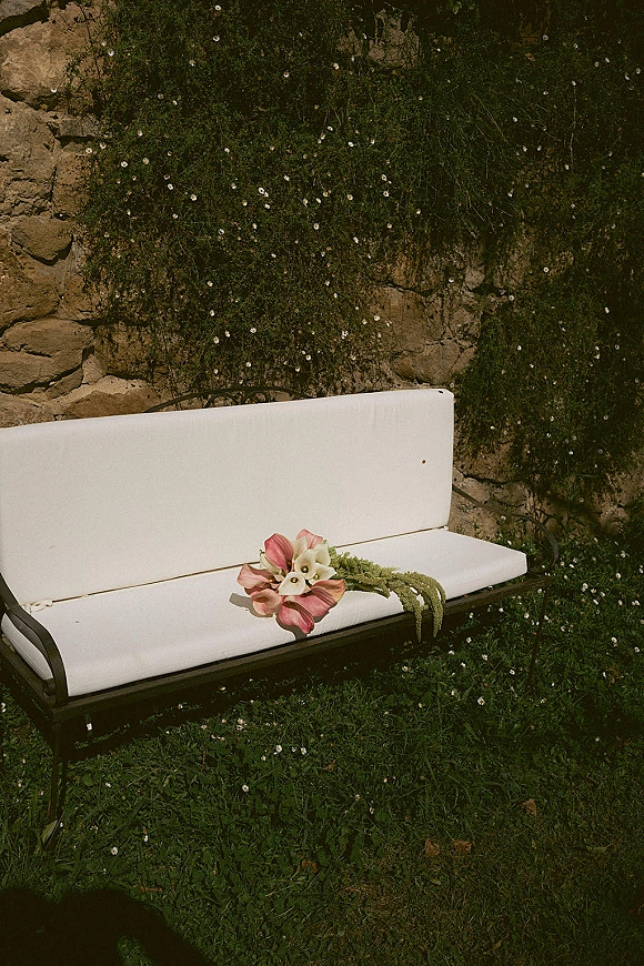 Wedding bouquet with calla lily bouquet, pink blooms and trailing greenery resting on a white outdoor sofa by an ivy stone wall