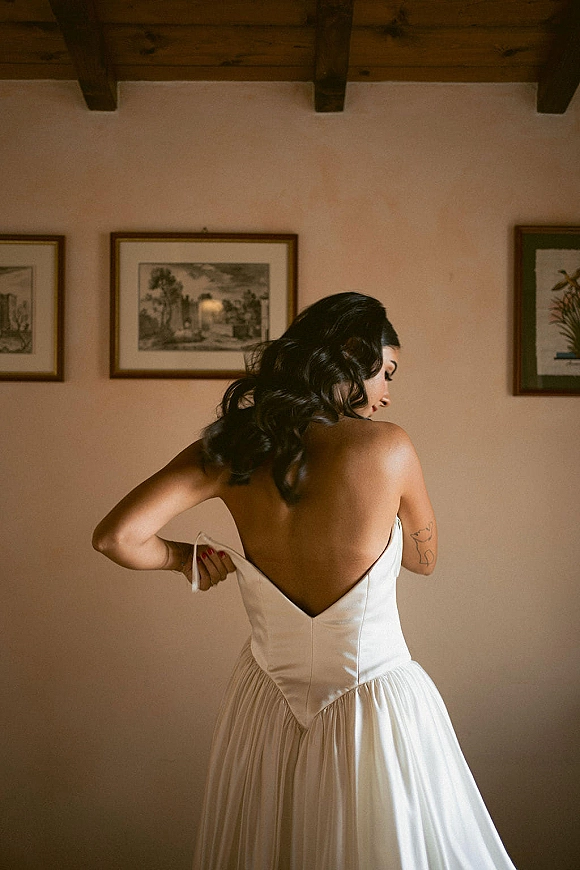 Bridal portrait of a bride in a strapless low back gown looking over her shoulder, showing arm tattoos, framed art and beams behind